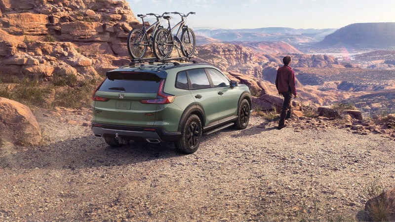 Green 2027 Honda CR-V with two mountain bikes on its roof rack, parked on a rocky canyon overlook as a man takes in the view.