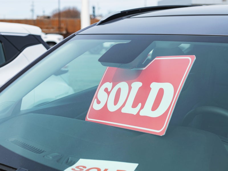 Red 'SOLD' sign displayed inside the windshield of a parked car, indicating the vehicle's purchase, set against a dealership background.